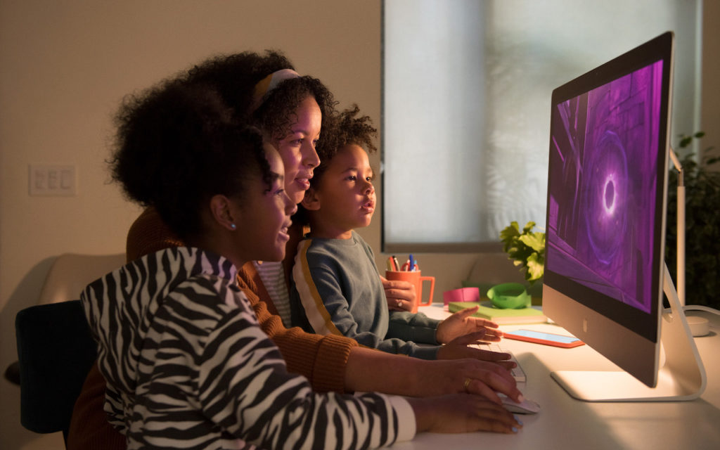 A woman and two young children sit closely together at a desk, looking intently at a large computer monitor displaying a purple screen in a warmly lit room.
