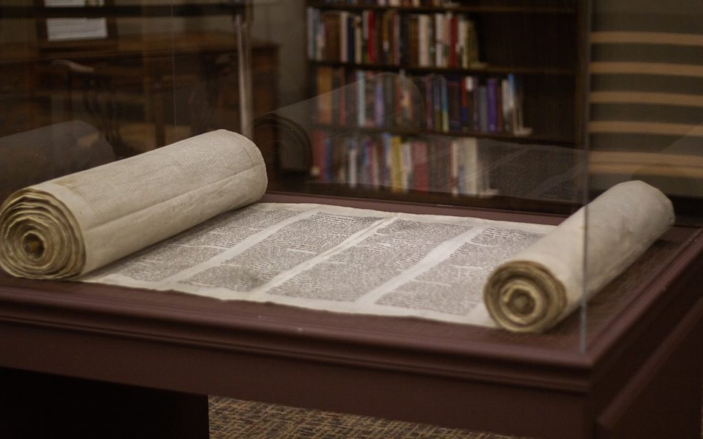 A large, ancient scroll with handwritten text is displayed in a glass case on a wooden stand, with a bookshelf and chair visible in the background.