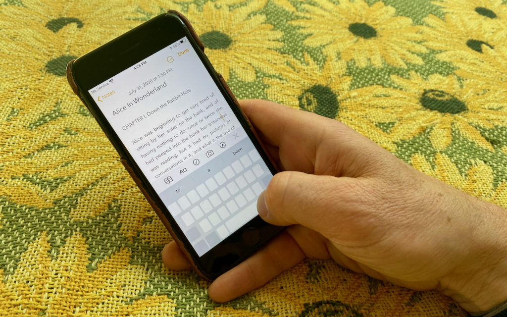 A hand holds a smartphone displaying text from Alice in Wonderland in the Notes app. The phone rests on a yellow tablecloth with a sunflower pattern.