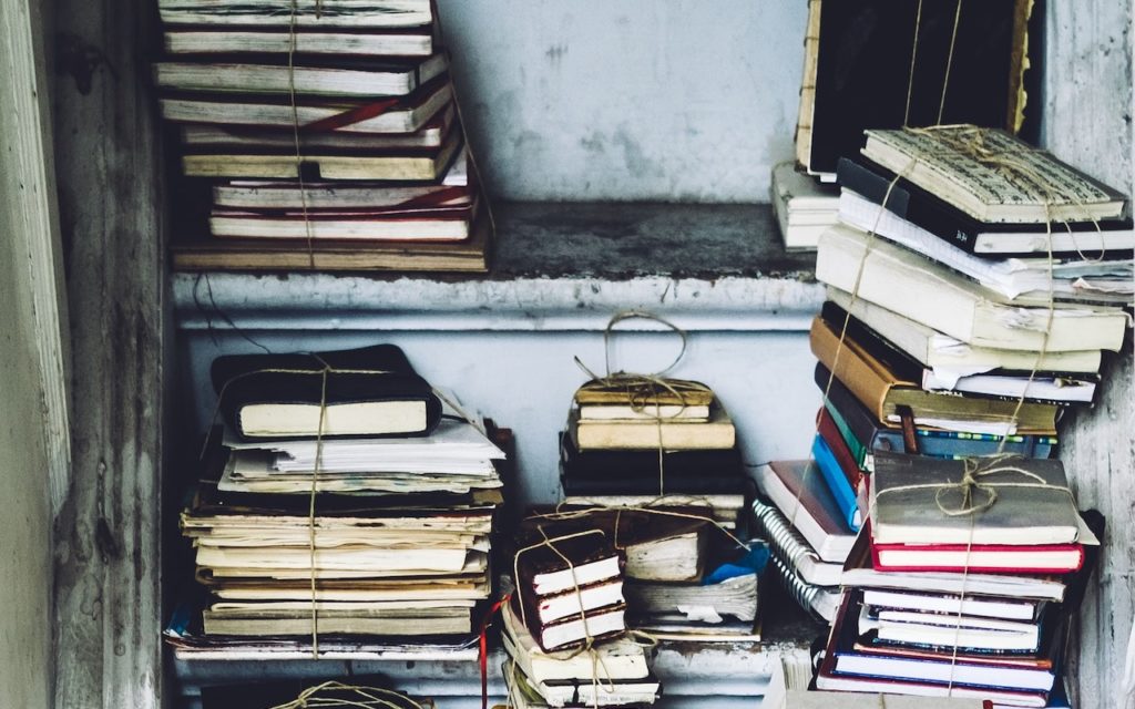 Stacks of old books and notebooks, some tied with string, are arranged haphazardly on worn shelves and surfaces against a faded wall, creating a cluttered but nostalgic atmosphere.