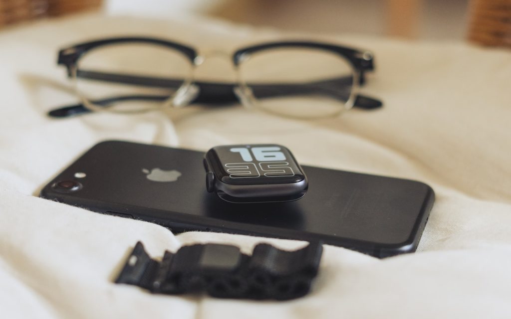 A black smartwatch with a digital display showing 16:35 rests on a black smartphone. A pair of eyeglasses and a detached black watch strap are placed on a light-colored fabric surface.