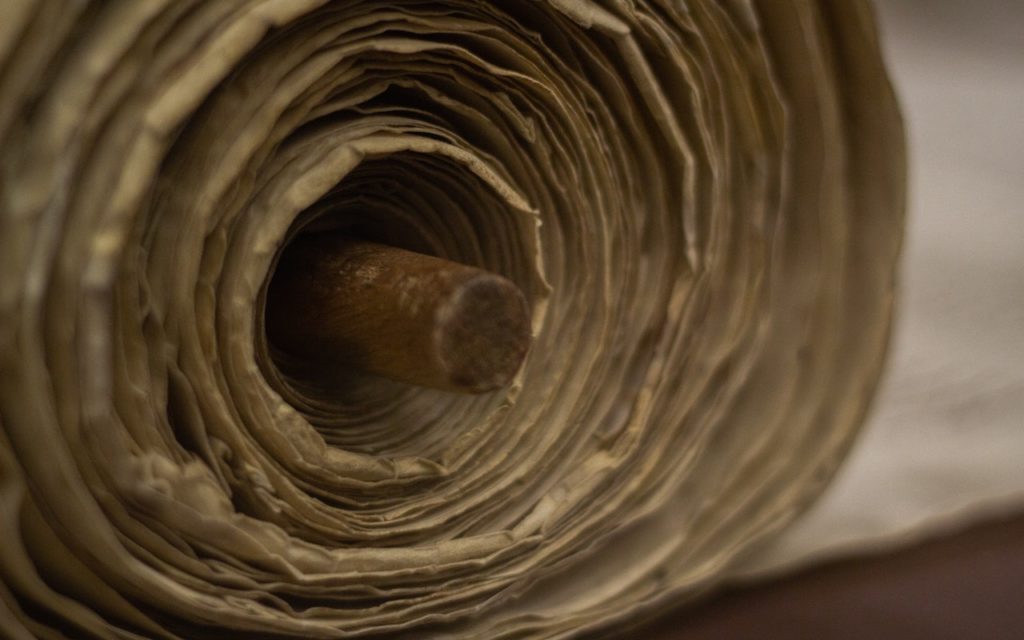 Close-up of the end of a large roll of thick, slightly wrinkled beige paper wound around a wooden rod, with layers of paper creating a spiral pattern.