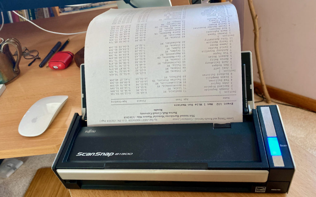 A Fujitsu ScanSnap iX500 scanner sits on a wooden desk, scanning a printed sheet with small, dense text. A computer mouse, pen, and red case are also visible on the desk nearby.