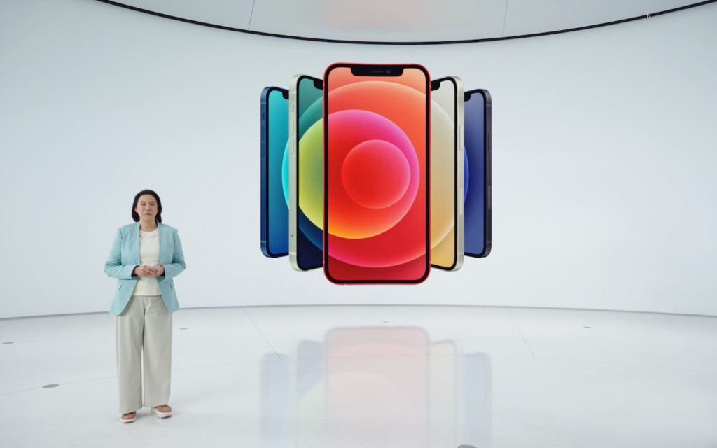 A woman in a light blue blazer stands in a white modern room next to a large digital display showing several colorful smartphones arranged in a semicircle.