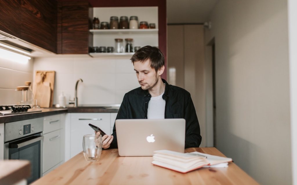 A man sits at a kitchen table with a laptop, glass mug, and open notebook, looking at his smartphone. Shelves with jars and a modern kitchen are visible in the background.