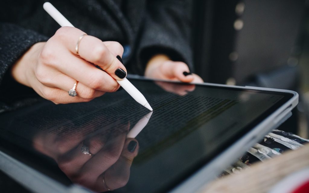 A person with black nail polish writes on a tablet with a stylus pen. The screen displays highlighted text, and the person is wearing a dark coat and a ring on their finger.