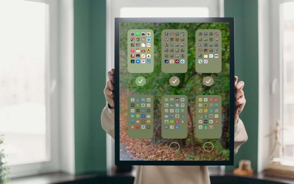 A person holds a large framed picture showing a smartphone screen with several app folders and icons, with a blurred outdoor background visible through the glass. The persons face is not visible.