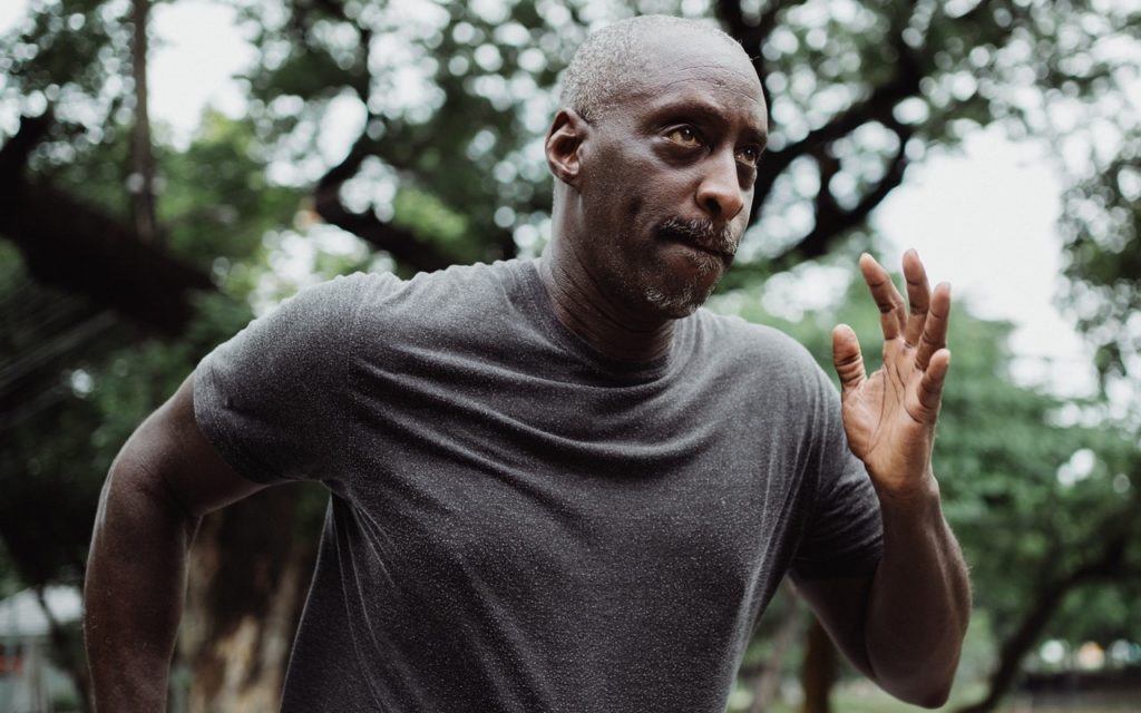 An older man wearing a gray t-shirt jogs outdoors, surrounded by trees. He appears focused and determined, with his arm raised mid-stride and greenery blurred in the background.