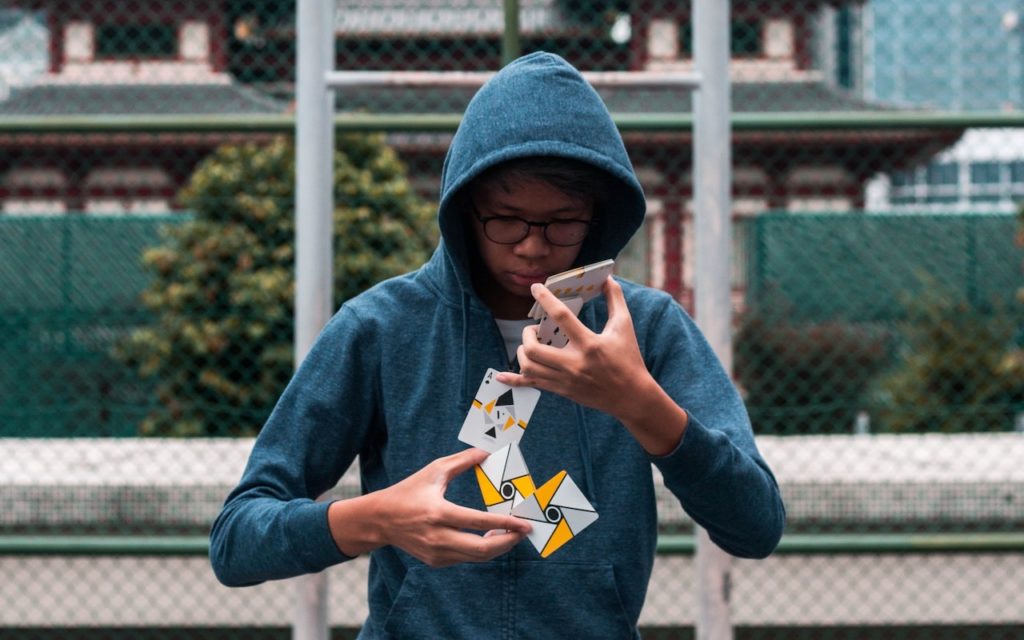 A person in a blue hoodie and glasses skillfully performs card tricks outdoors, holding and spreading several playing cards with both hands. A green fence and buildings are visible in the background.