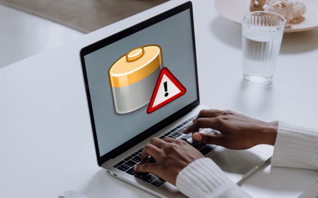 A person types on a laptop displaying a large battery icon with an exclamation mark warning symbol on the screen, indicating a low battery alert. A glass of water and a bowl are on the white table nearby.
