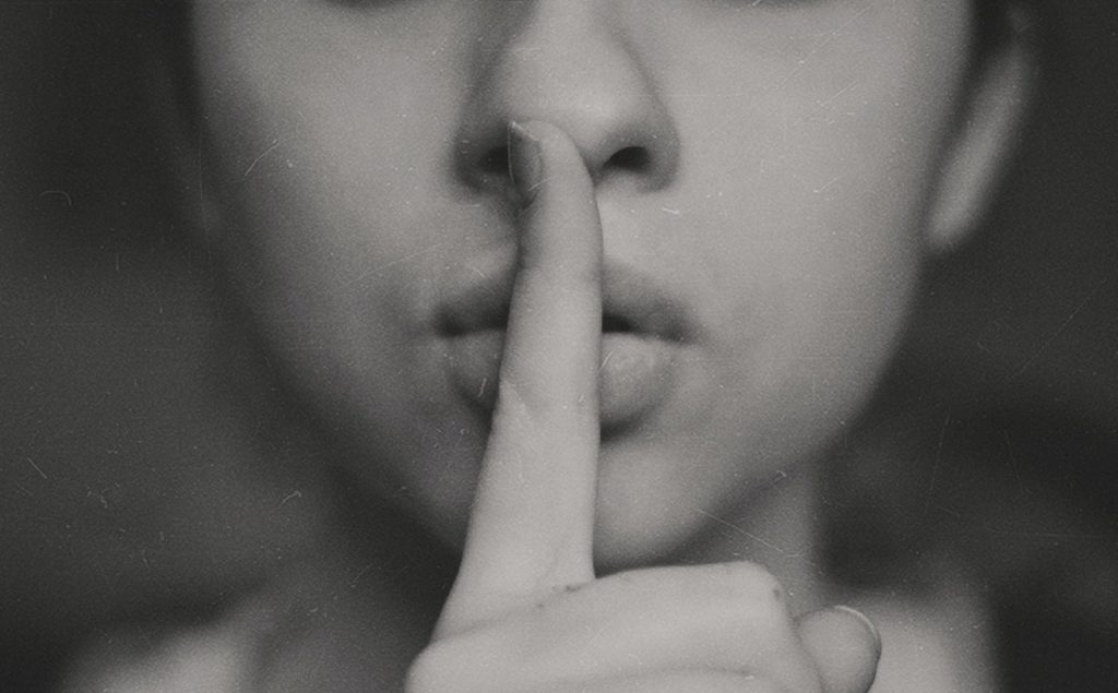 A grayscale close-up of a person’s face, with their index finger held vertically over their lips, signaling for silence or secrecy. The background is blurred and the image has a soft, grainy texture.