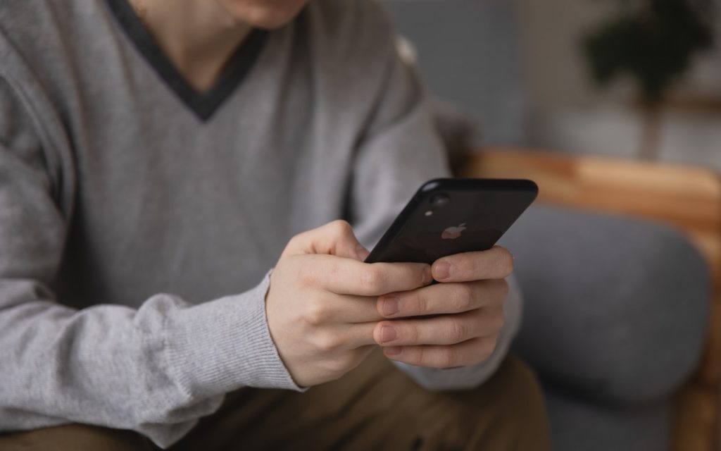 A person wearing a gray sweater is sitting and holding a black smartphone with both hands, looking at the screen. The background is softly blurred, focusing on the phone and hands.