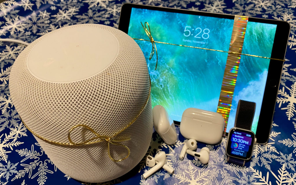 A HomePod, AirPods, Apple Watch, and iPad decorated with gold ribbons are displayed on a snowflake-patterned blue background, suggesting tech-themed holiday gifts.