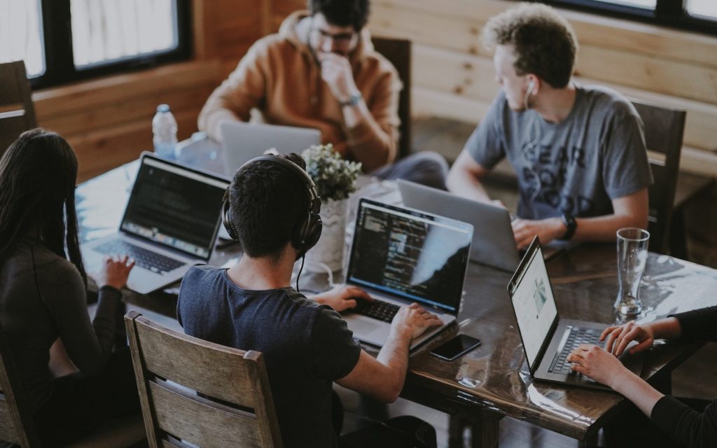 Five people sit around a wooden table working on laptops in a bright room. One person wears headphones, and there are drinks and a small plant on the table. They seem to be collaborating or working together.