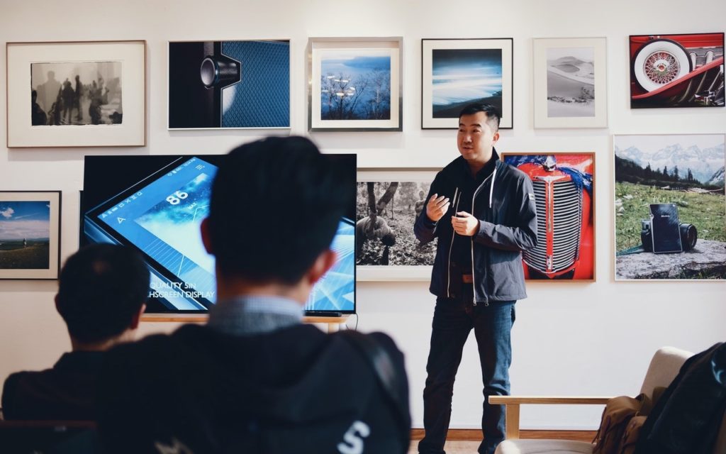A man stands and gives a presentation to an audience in a room decorated with framed photos and artwork. A large screen displaying a digital image is beside him. Two people sit and listen in the foreground.