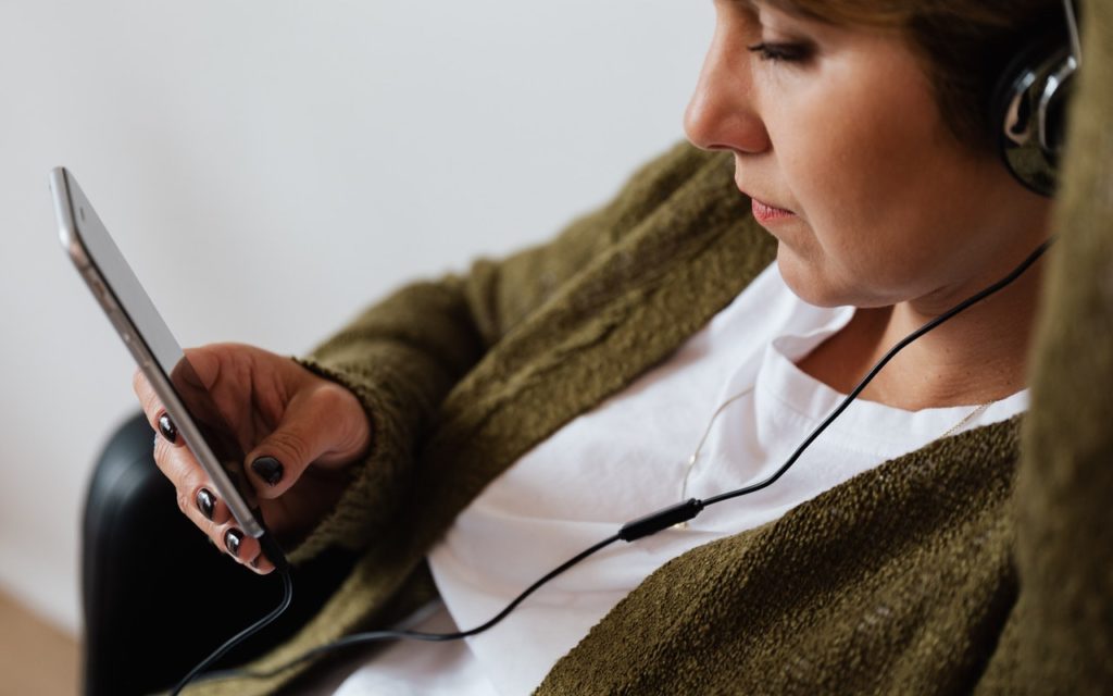 A person wearing headphones and a green sweater sits and looks at a smartphone, holding it in one hand. Their attention is focused on the screen.