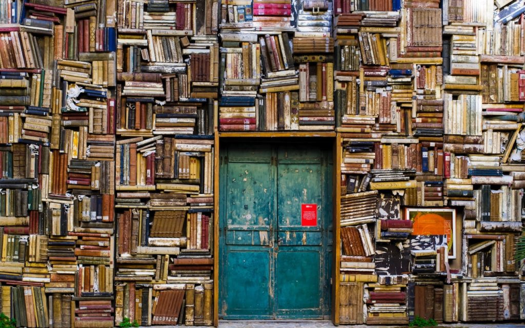A green double door set in a wall completely covered with stacked and crammed old books; some books are open, leaning, or unevenly arranged, creating a colorful, textured backdrop.