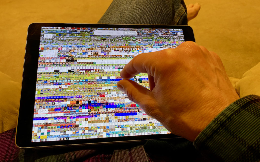 A person’s hand interacts with a tablet displaying a grid of many small, colorful photo thumbnails. The person is seated, wearing jeans and a striped shirt, with one bare foot visible in the background.