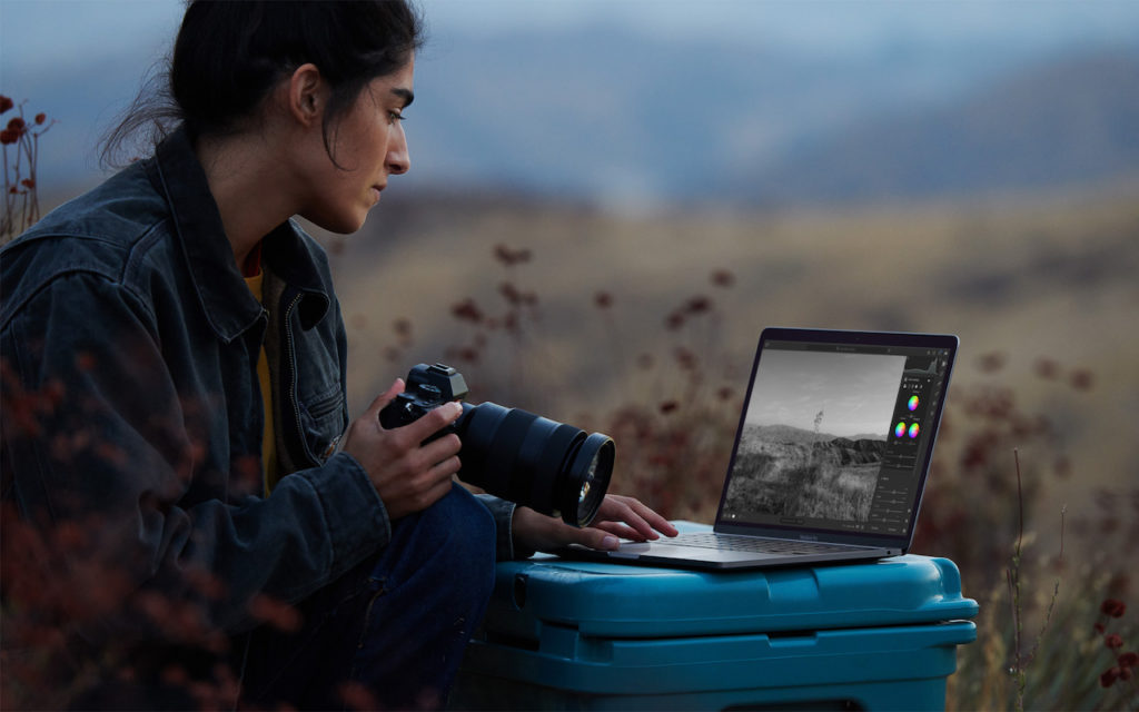A person holding a camera sits outdoors on a blue case, editing a black-and-white photo on a laptop. The background shows blurred hills and dry grass, suggesting a natural, rural setting.