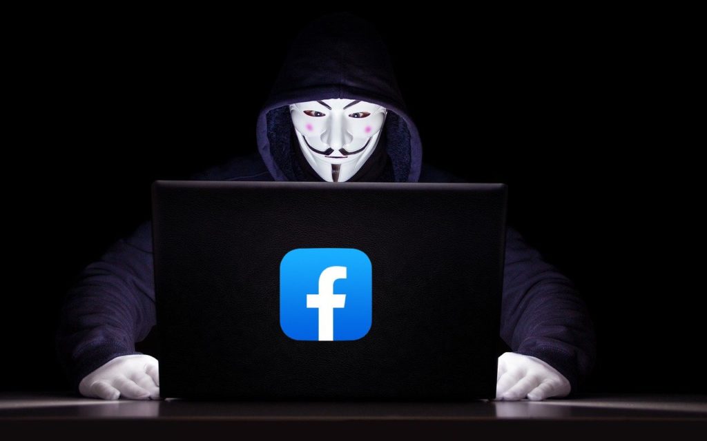 A person wearing a Guy Fawkes mask and a dark hoodie sits in front of a laptop with a large blue Facebook logo on it, against a black background.