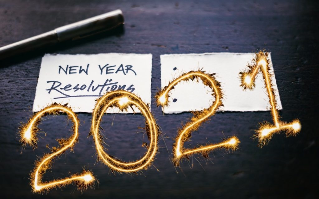 A pen and two paper notes on a dark surface; one note says New Year Resolutions, and the other shows 2021 written with bright sparkler light.