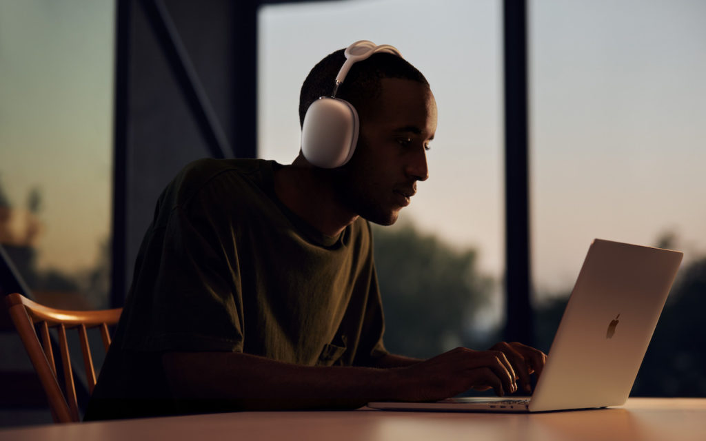 A person wearing large white headphones sits at a table, focused on typing on a laptop. The background shows large windows with a sunset view, casting a warm light into the room.