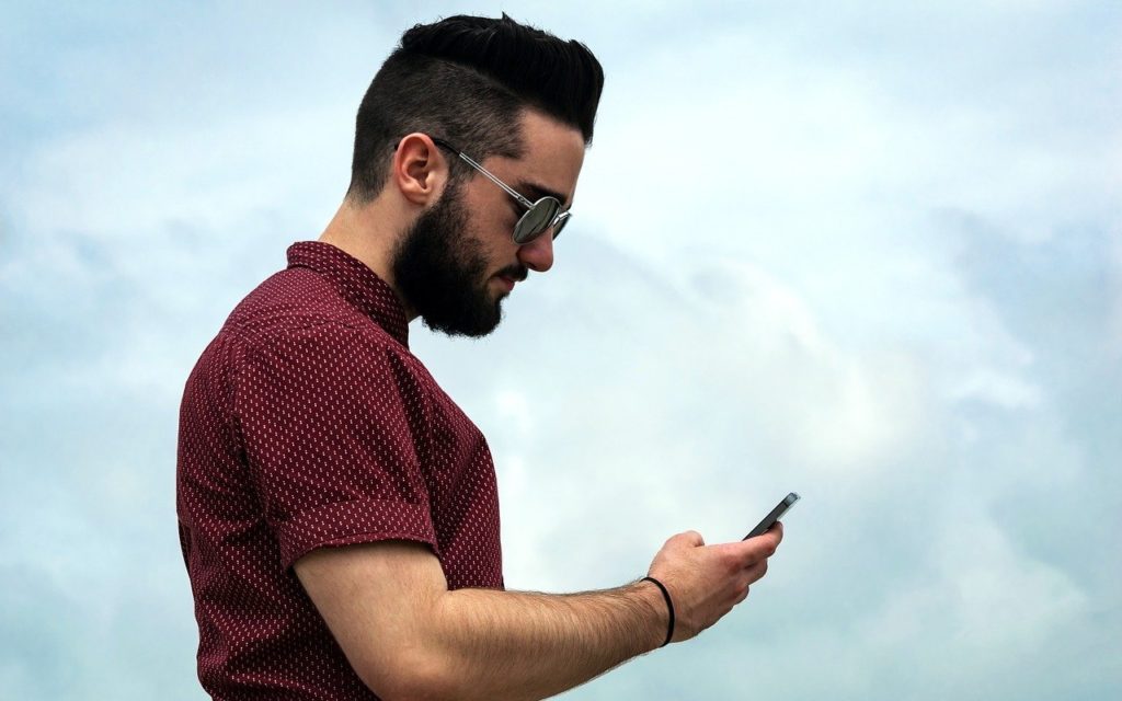A bearded man wearing sunglasses and a red patterned shirt looks down at his smartphone while standing outside against a cloudy sky.