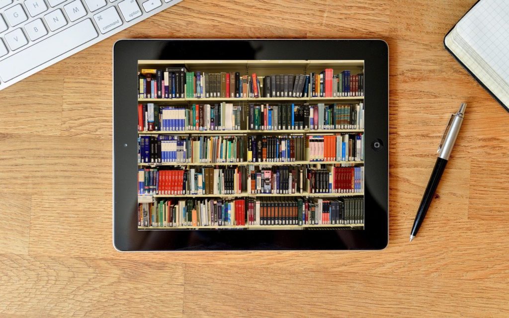 A tablet on a wooden desk displays an image of bookshelves filled with books. Nearby are a keyboard, an open notebook, and a pen.