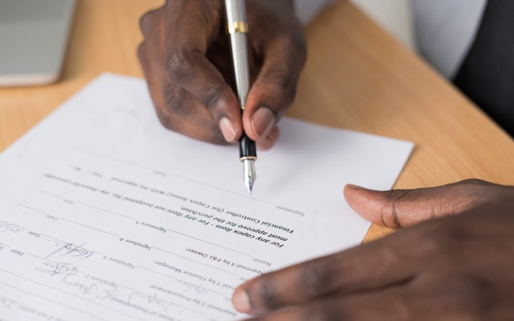 A person with dark skin signs a document with a silver pen while sitting at a wooden desk. Only the hands and part of the paper are visible.