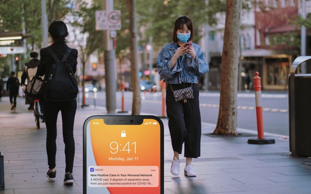 A large smartphone screen in the foreground shows a COVID-19 exposure alert. In the background, a woman wearing a face mask walks on a city street while looking at her phone, and another person walks away.