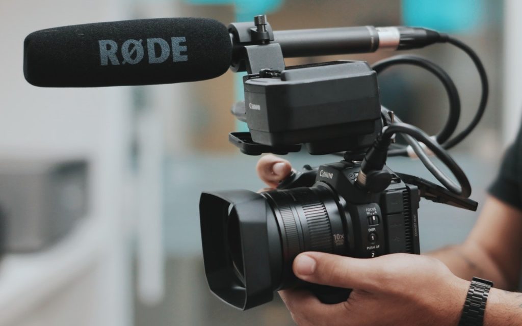 A person holds a professional Canon camera with a large lens and an attached RODE microphone, preparing for video recording. The background is blurred.