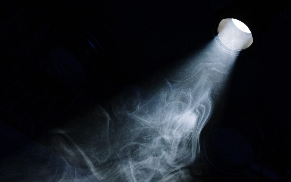 A beam of light from a flashlight shines through swirling smoke or mist in a dark environment, creating a dramatic and mysterious effect.