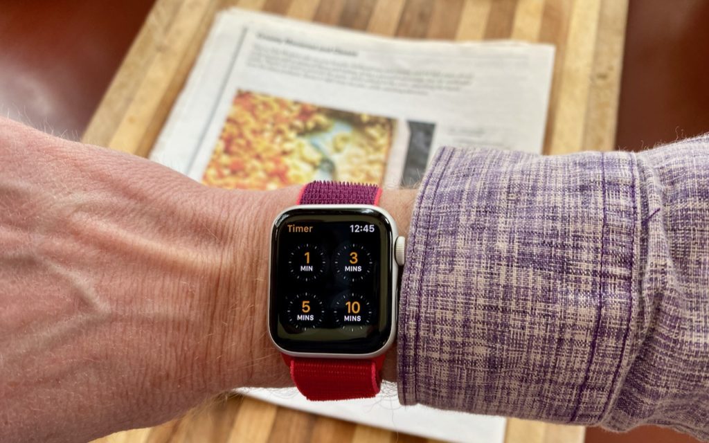 A person wearing a red-banded smartwatch with a timer app open, showing 1, 3, 5, and 10 minute options. In the background, a recipe page with a food image is seen on a wooden surface.