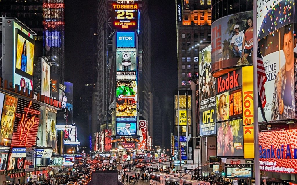 A lively night scene in Times Square, New York City, filled with bright electronic billboards, colorful advertisements, and crowds of people and vehicles.