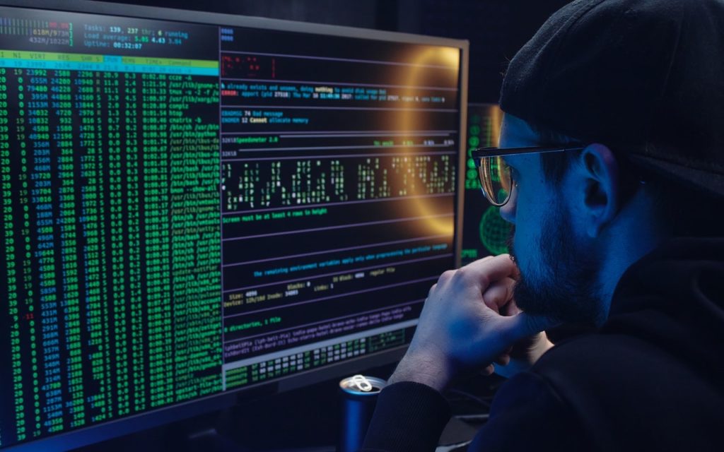 A person wearing a cap and glasses sits in front of computer monitors displaying lines of code in green and blue, focusing intently on the screens in a dimly lit room.