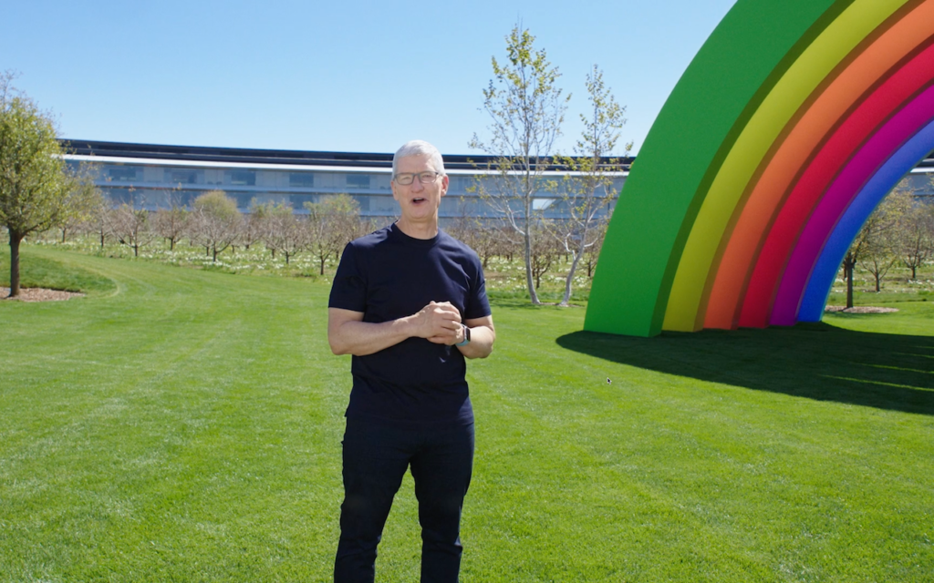 A man in a black shirt stands on green grass near a large rainbow-colored arch, with trees and a modern building in the background on a sunny day.