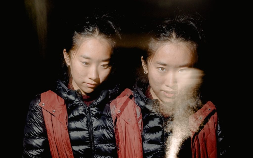 A woman wearing a black puffer jacket and maroon vest appears twice in the image, side by side, with a blurred light streak crossing in front of her, creating a double exposure effect against a dark background.