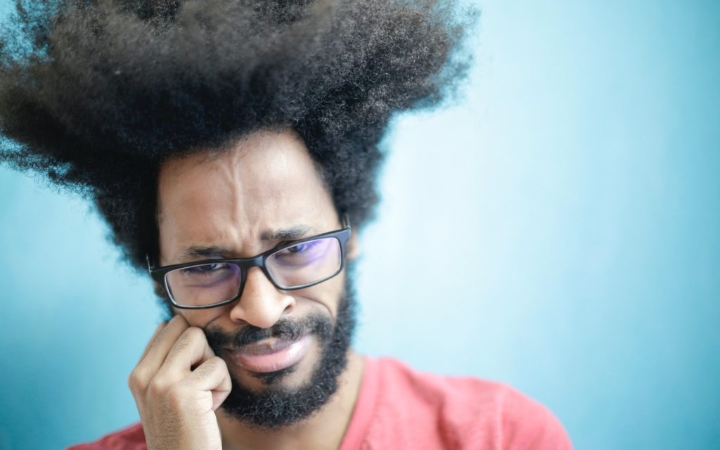 A man with a beard and glasses stands against a blue background, frowning with a puzzled expression while touching his cheek with one hand. His hair is voluminous and stands out in all directions.