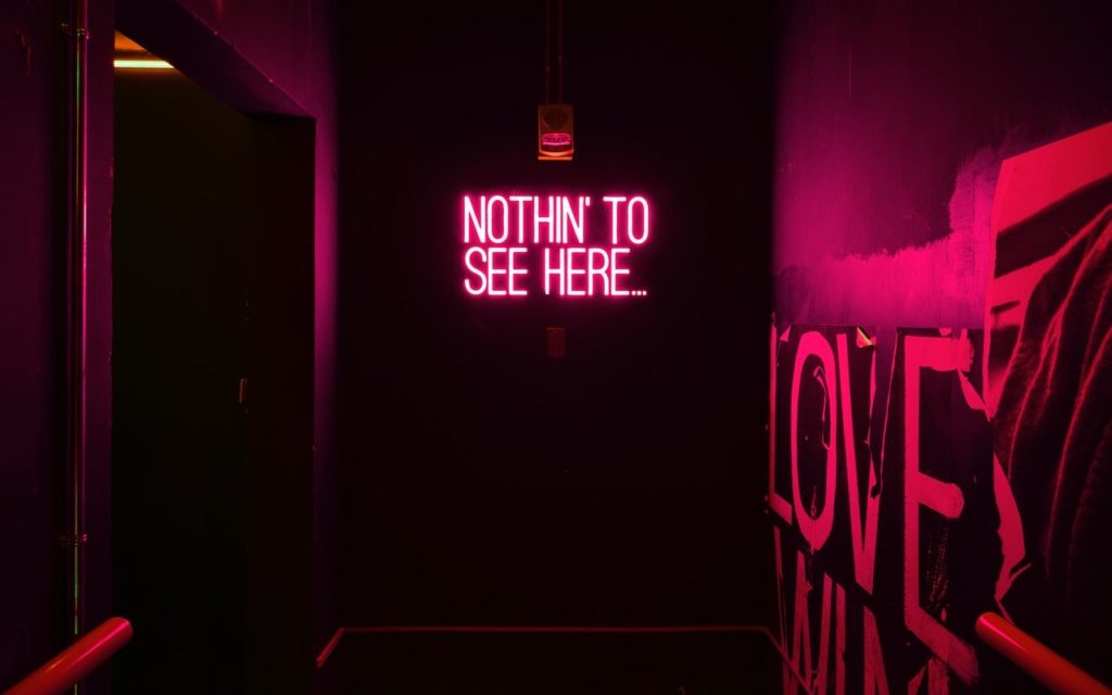 A dimly lit hallway with pink-purple neon lighting features a neon sign on the back wall that reads, “NOTHIN’ TO SEE HERE…” and part of a wall mural with the word “LOVE” partially visible on the right.