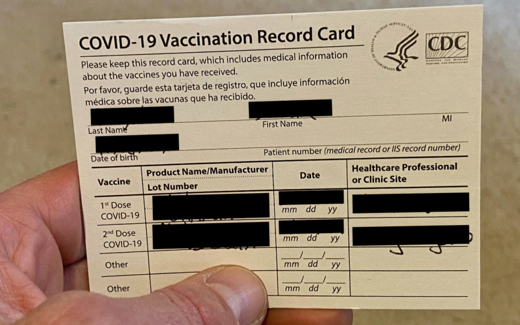 A hand holds a COVID-19 Vaccination Record Card showing sections for name, date of birth, vaccine manufacturer, dates of doses, and healthcare provider information, with personal details blacked out.