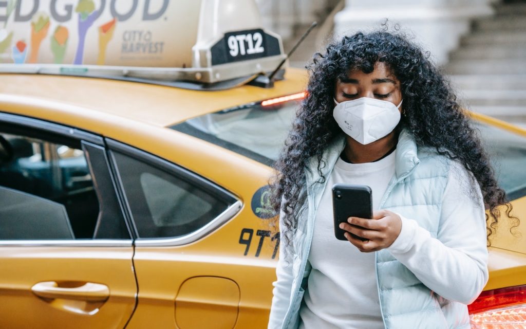 A woman with long curly hair, wearing a face mask and light jacket, stands next to a yellow taxi while looking at her smartphone.