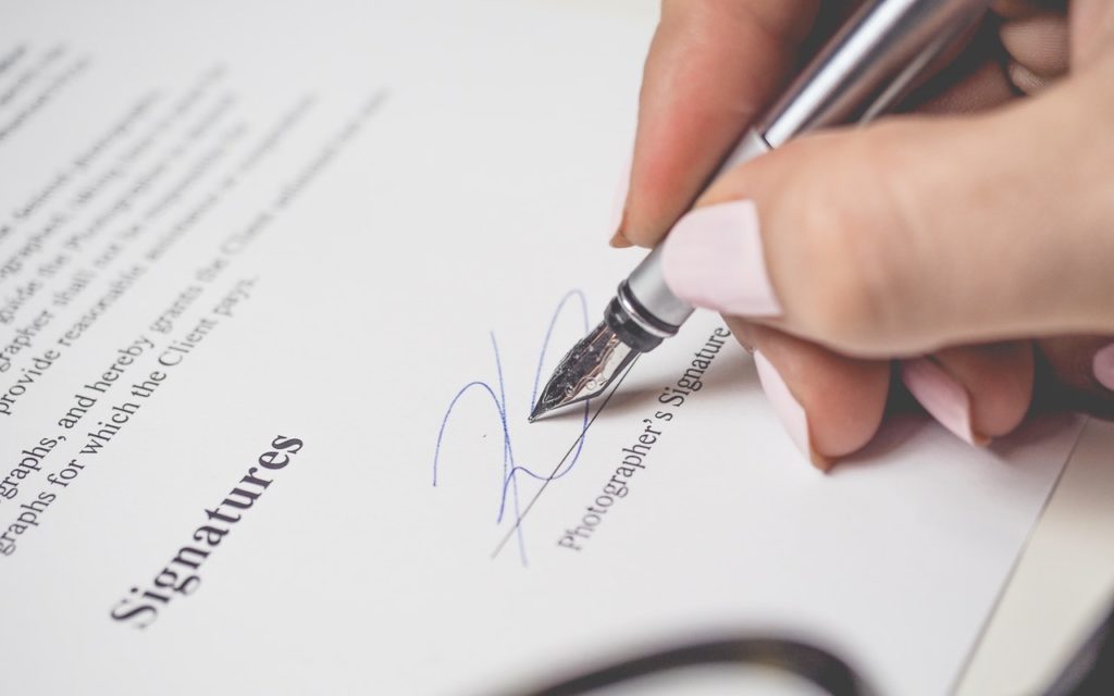 A person with light pink nail polish signs a document labeled Signatures with a silver pen. The hand is close to the paper, focusing on the act of signing.