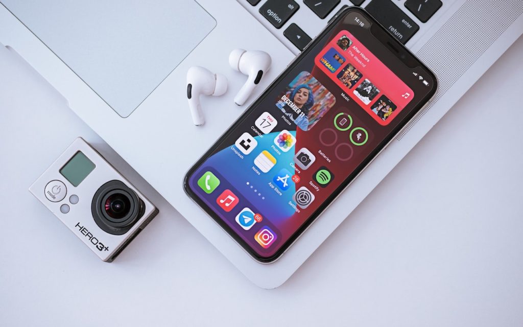 A GoPro camera, a pair of white wireless earbuds, and a smartphone with colorful app icons and music widgets on its screen rest on a silver laptop keyboard.