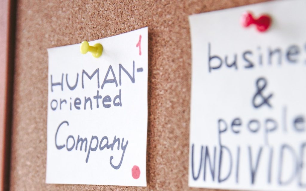 A handwritten note pinned to a corkboard reads HUMAN-oriented Company with another blurred note nearby mentioning business & people UNDIVI.... Yellow and red push pins secure the notes.