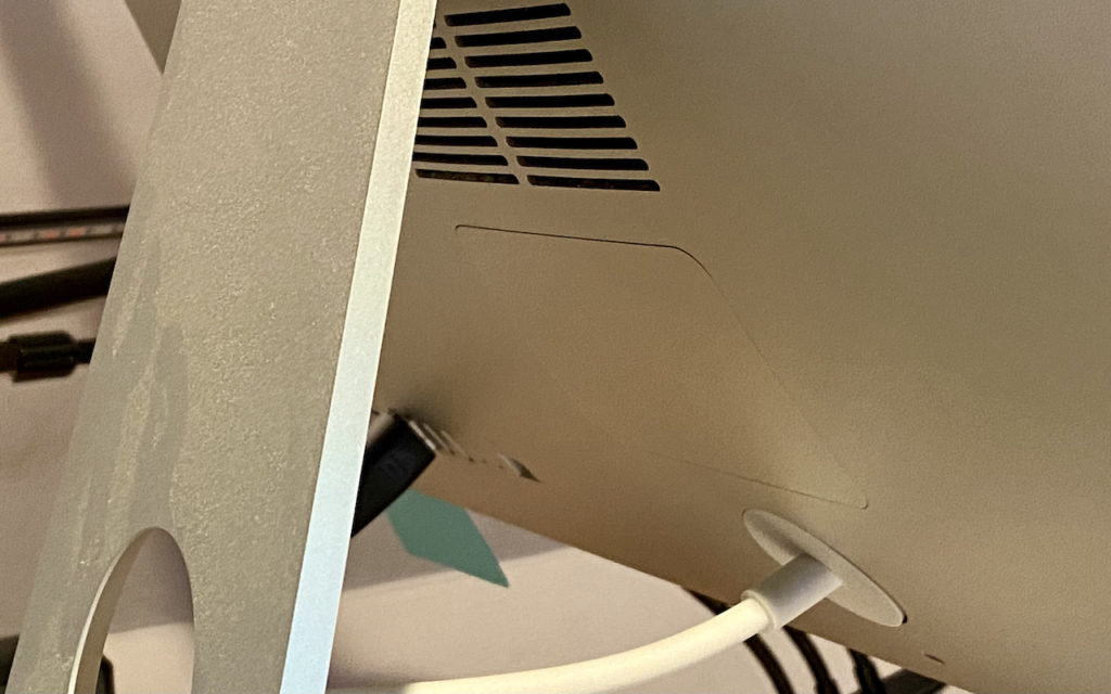Close-up of the back of a computer monitor showing its ventilation slots, stand, ports with cables plugged in, and a network of cables beneath.