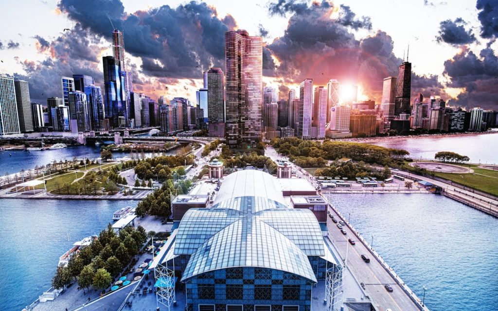 Aerial view of a modern city skyline at sunset with tall glass buildings, a domed pavilion, tree-lined paths, and water on both sides reflecting the colorful sky and clouds.