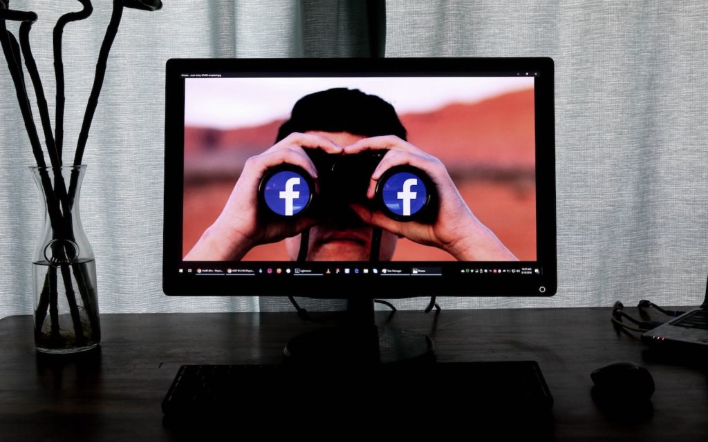A computer monitor displays an image of a person looking through binoculars with Facebook logos on the lenses. The monitor sits on a desk with a keyboard and a vase in the background.