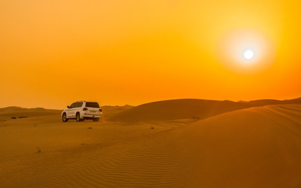 A white SUV drives across sandy dunes under a bright, orange sunset in a vast desert landscape. The sun glows high in the sky, casting warm light over the rippled sand.