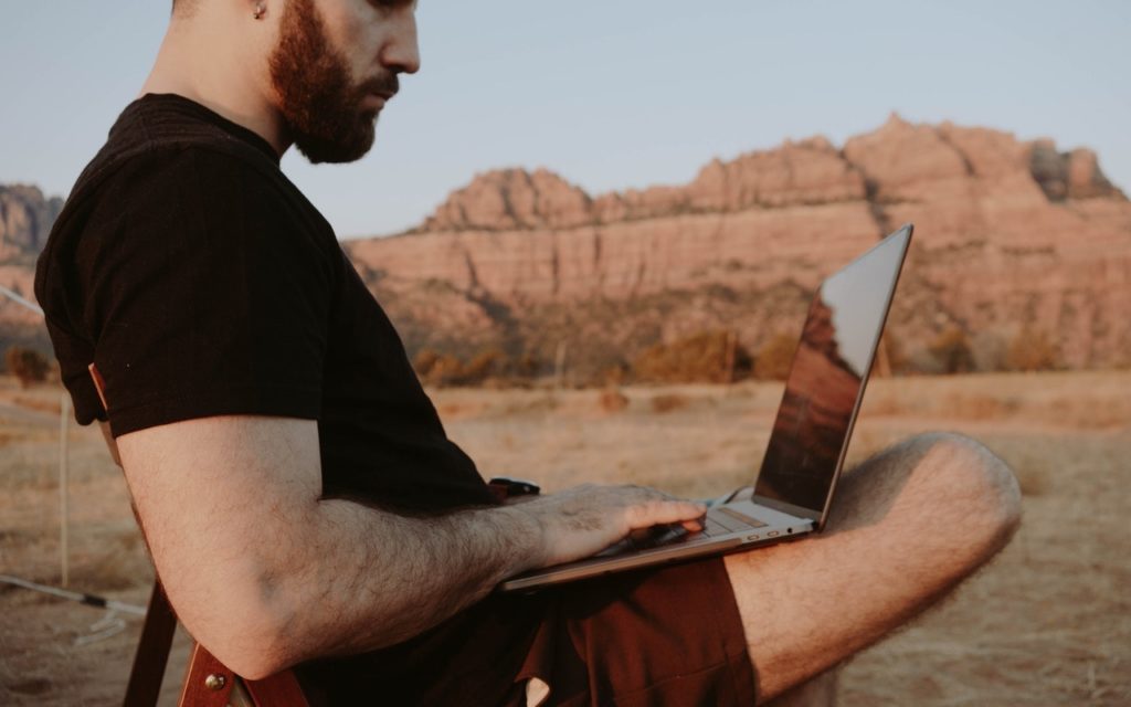 A man with a beard sits outdoors on a chair, using a laptop on his lap. He is wearing a black t-shirt and shorts, with rocky mountains and dry grassland in the background under a clear sky.
