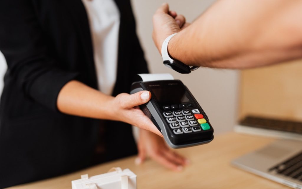 A person pays using a smartwatch by holding their wrist over a card reader held by another person, who is dressed in business attire at a checkout counter.
